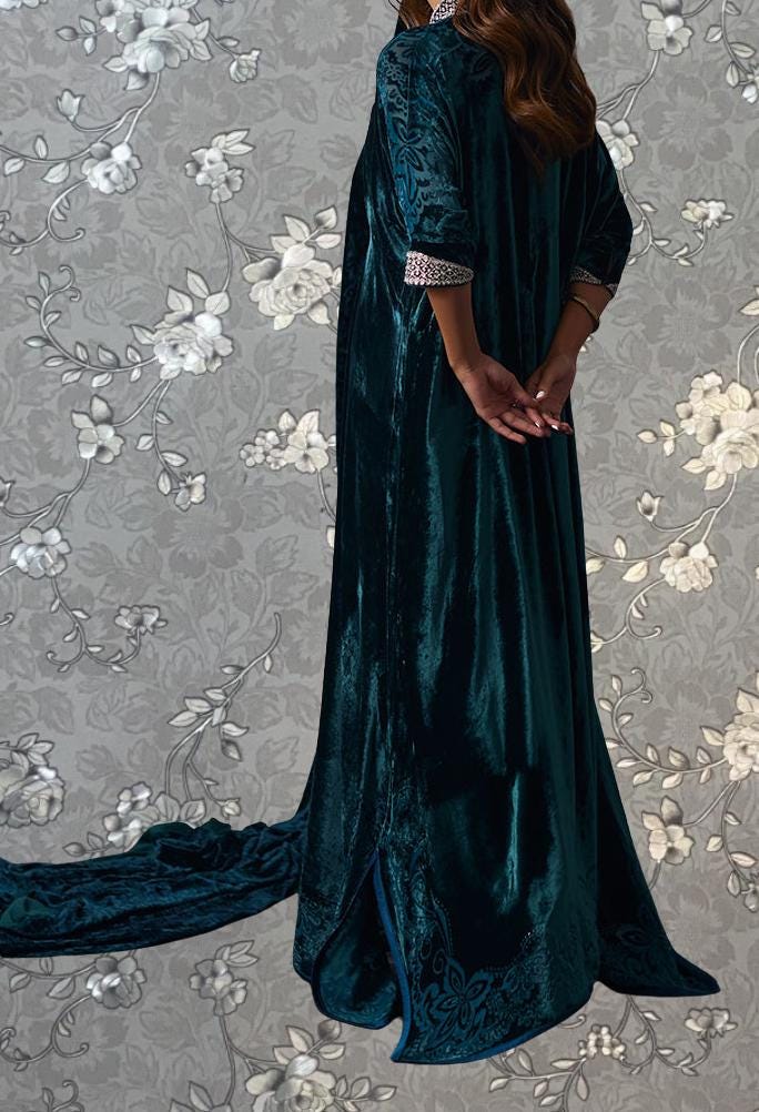 A woman wearing a long, dark green velvet dress with a floral pattern stands against a gray floral background.