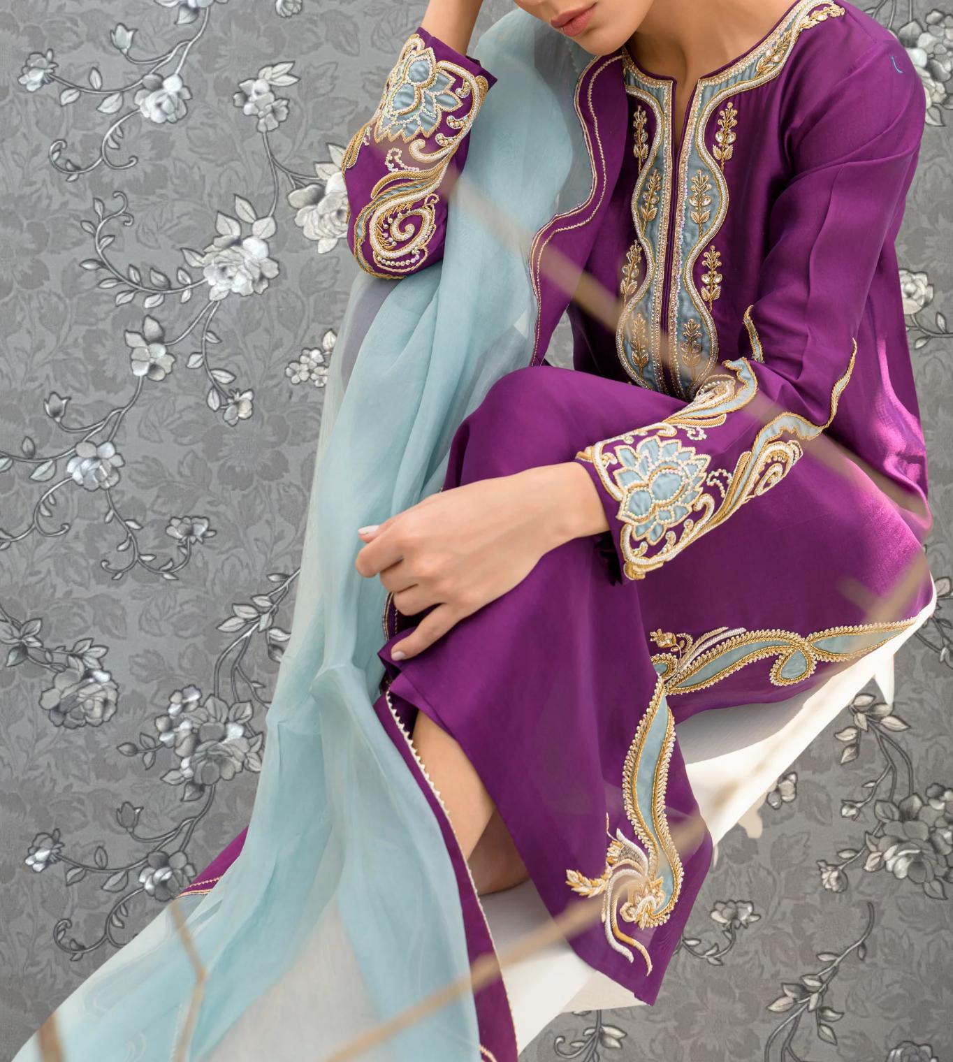 A person wearing a purple and gold dress with intricate embroidery is sitting on a chair against a gray floral background.