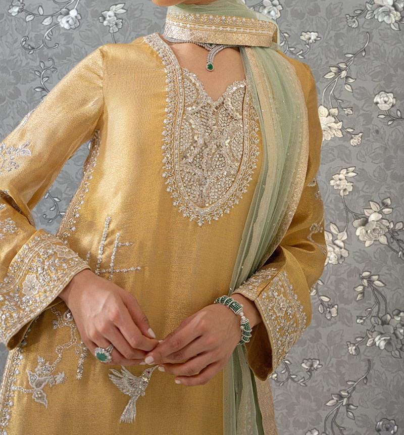 A woman wearing a gold and green dress with intricate embroidery and jewelry poses against a gray floral background.