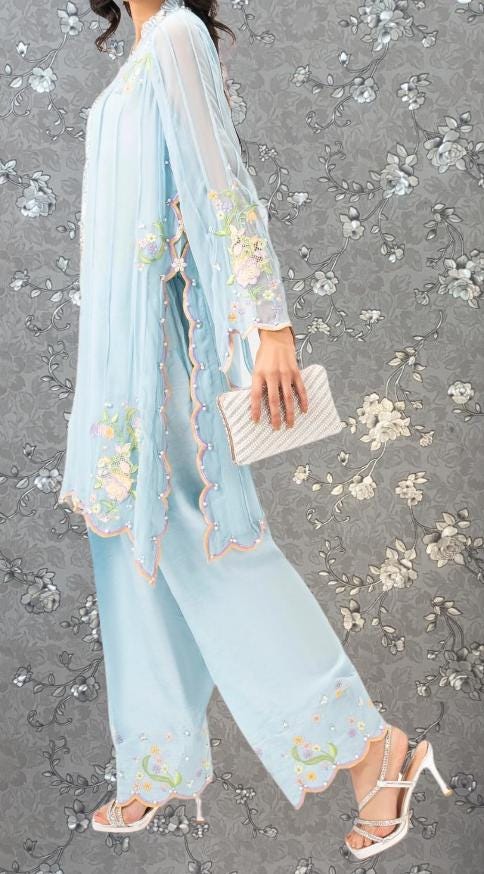 A woman wearing a light blue dress with floral embroidery and holding a small white clutch purse is standing against a gray floral background.