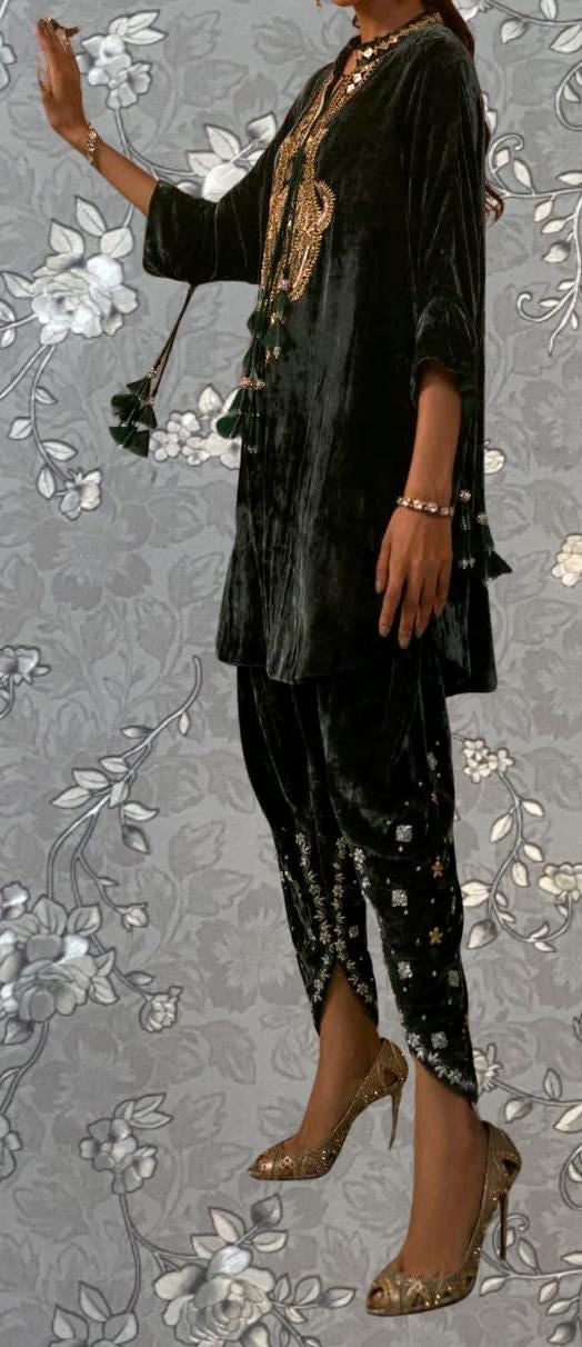 A person wearing a black velvet dress with gold embroidery and tassels, paired with gold high heels, stands against a gray floral background.