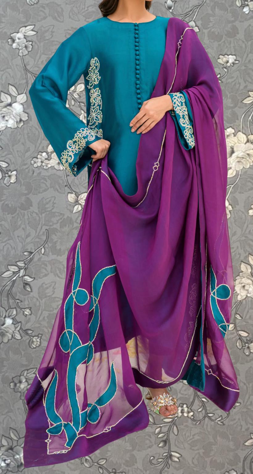 A woman wearing a teal blue dress with a purple shawl and a patterned skirt, standing against a gray floral background.