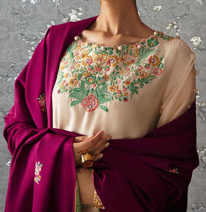 A woman wearing a pink shawl and a white blouse with a floral embroidered design, standing against a gray floral background.