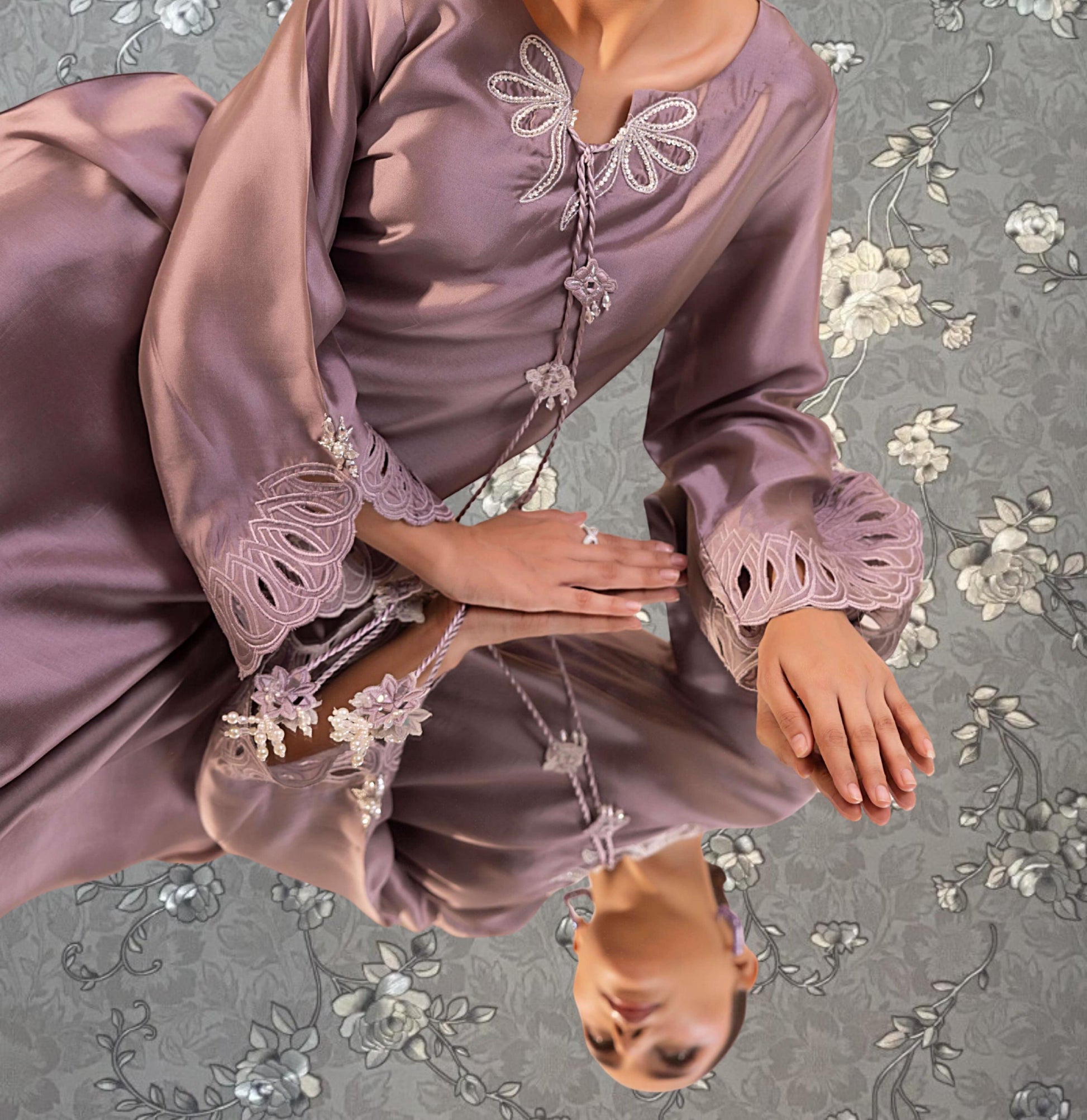 A woman wearing a long, purple dress with lace details and a floral pattern, posing with her hands on her lap.