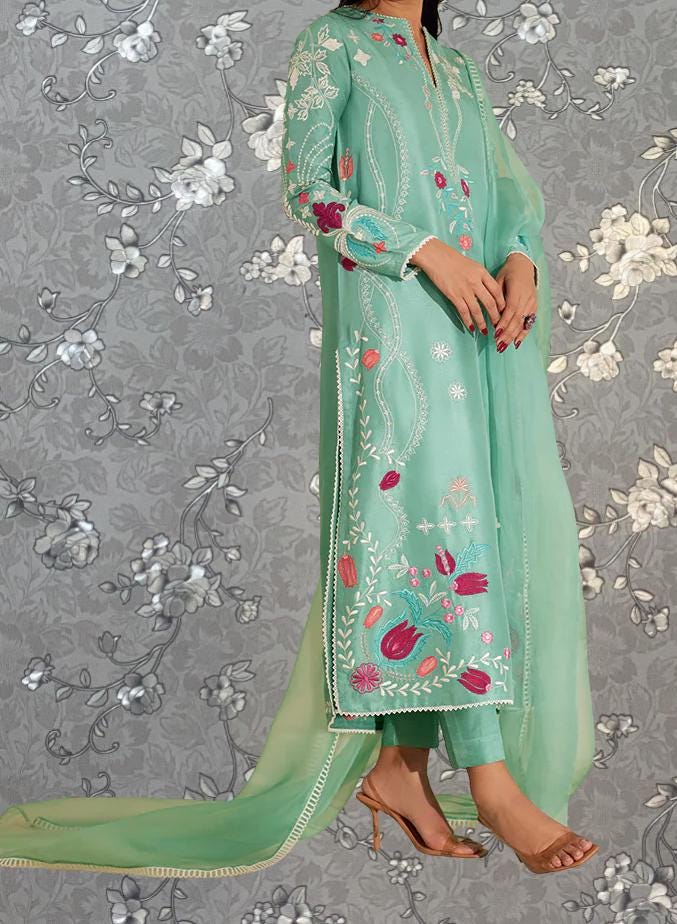 A woman wearing a long, mint green dress with intricate floral embroidery stands against a gray floral background.