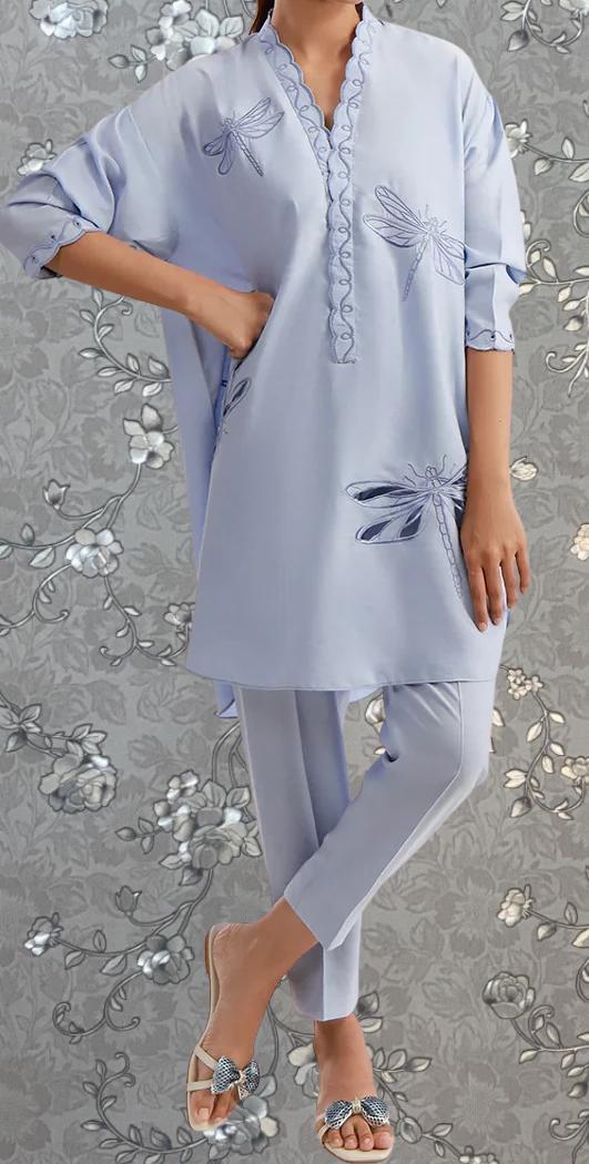 A person wearing a light blue dress with a dragonfly design on it, standing against a gray floral background.