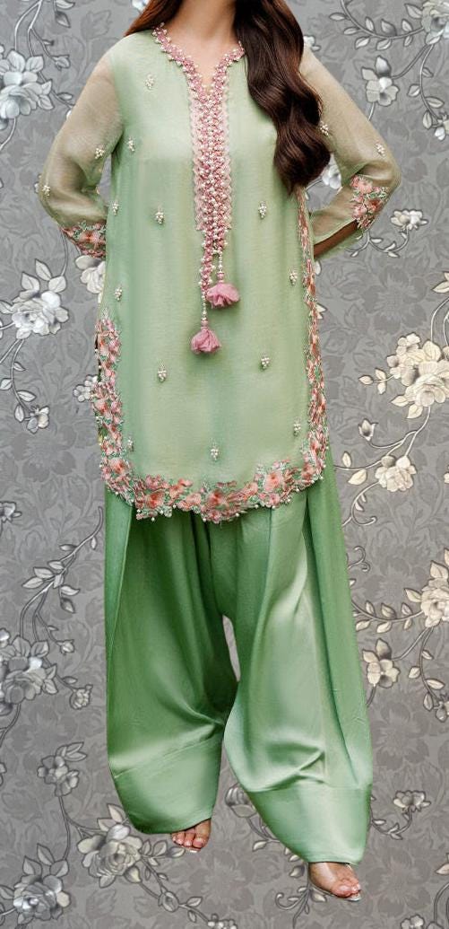 A woman wearing a green dress with pink accents and floral embroidery, standing against a gray floral background.