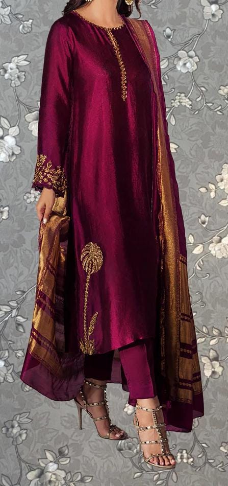 A woman wearing a long, burgundy-colored dress with gold embroidery and a matching gold and gold shawl. She is wearing strappy gold heels and has her hand in her pocket.