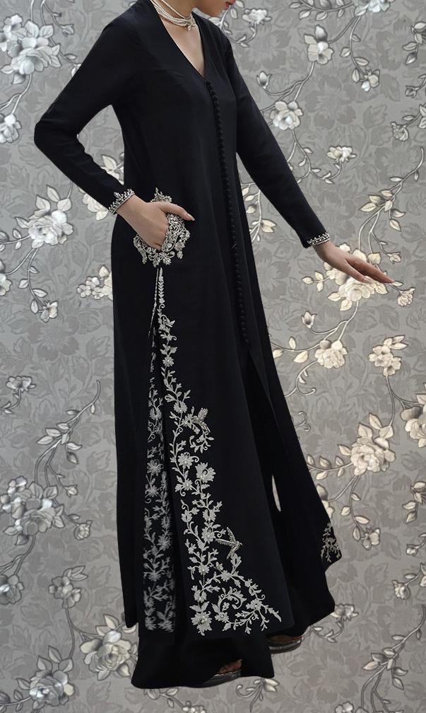 A person wearing a long black dress with intricate white floral embroidery stands against a gray floral background.