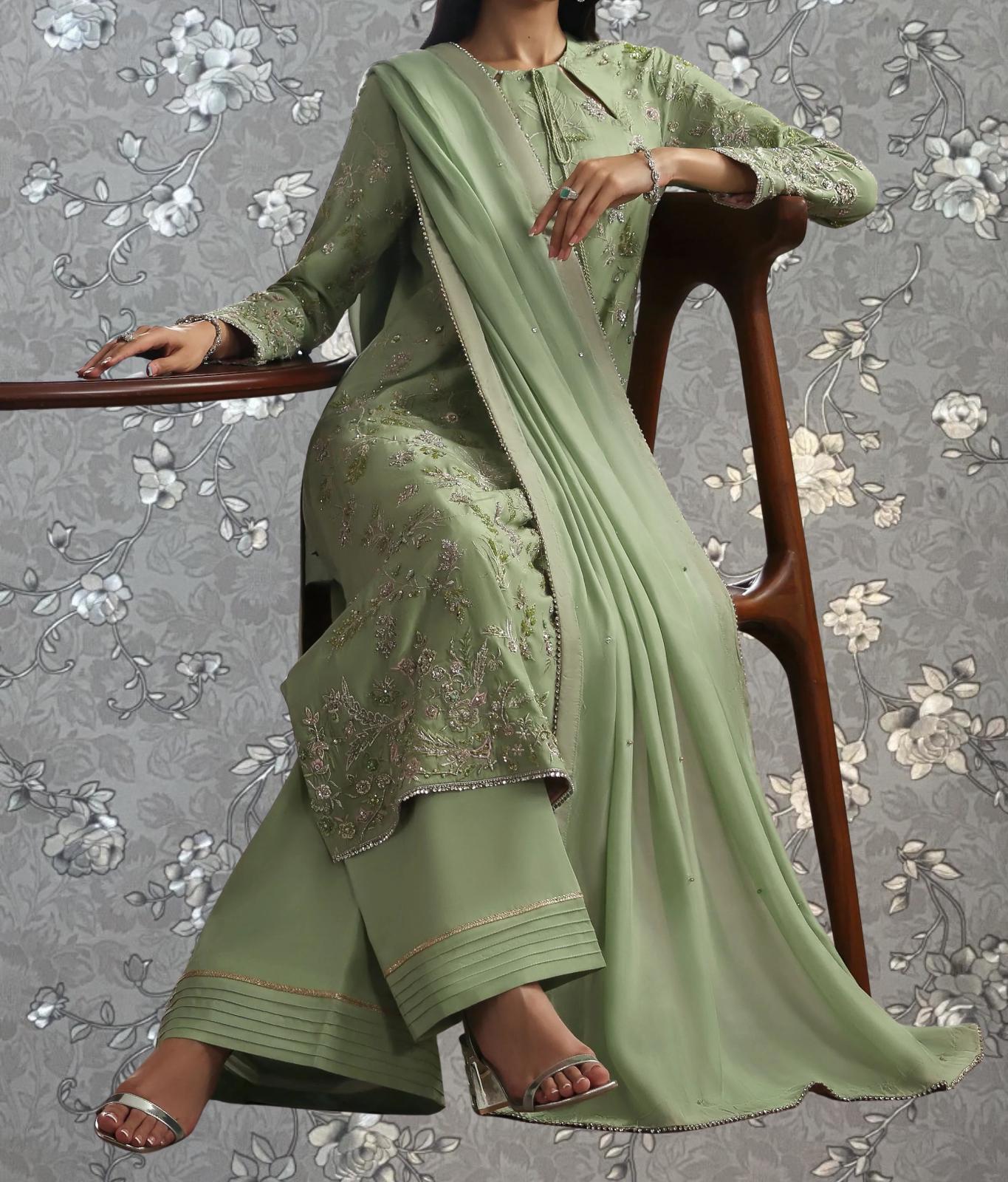 A woman wearing a green and gold dress with intricate embroidery, sitting on a wooden chair against a gray floral background.