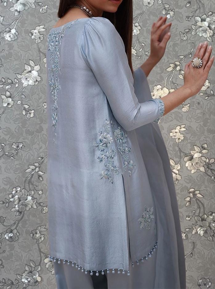 A woman wearing a light blue dress with intricate embroidery on the back stands against a floral patterned background.