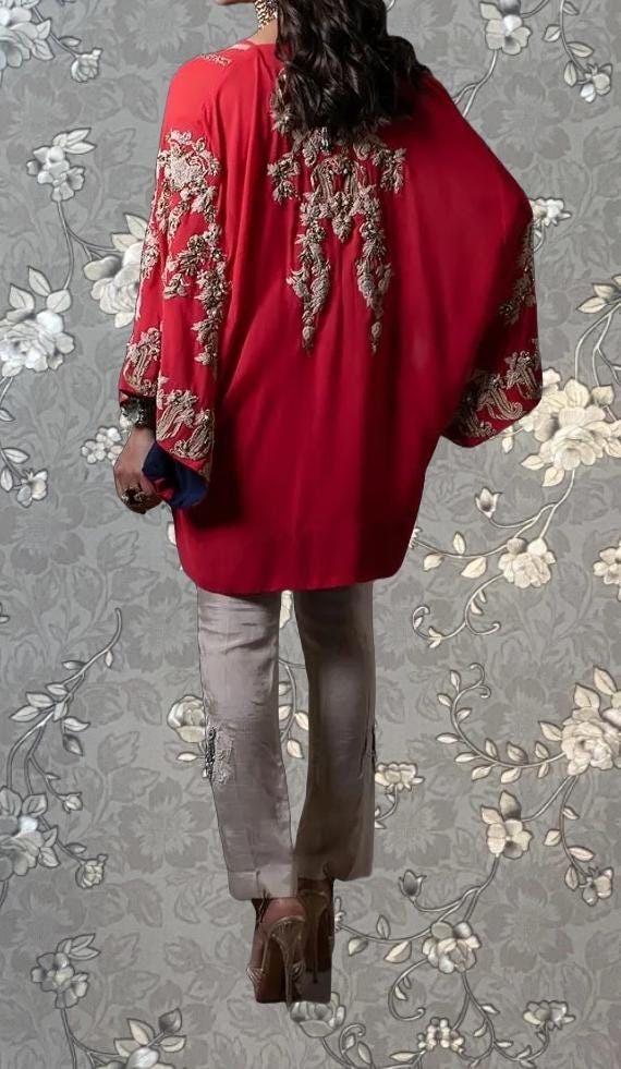 A person wearing a red kurta with intricate embroidery on the back, standing against a gray floral background.