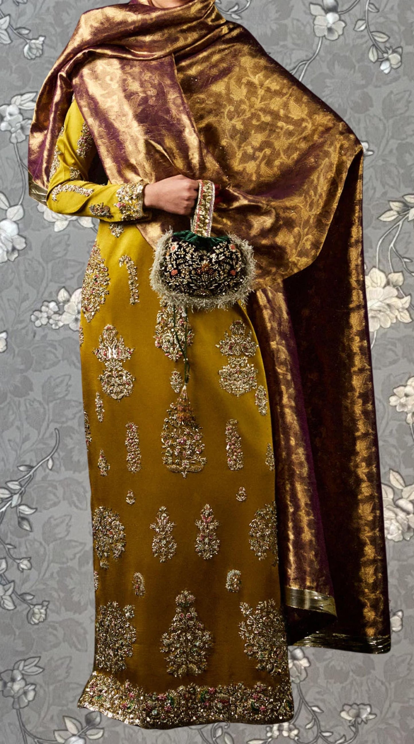 a person wearing a gold and yellow dress with intricate embroidery, holding a small purse, and standing against a gray floral background.