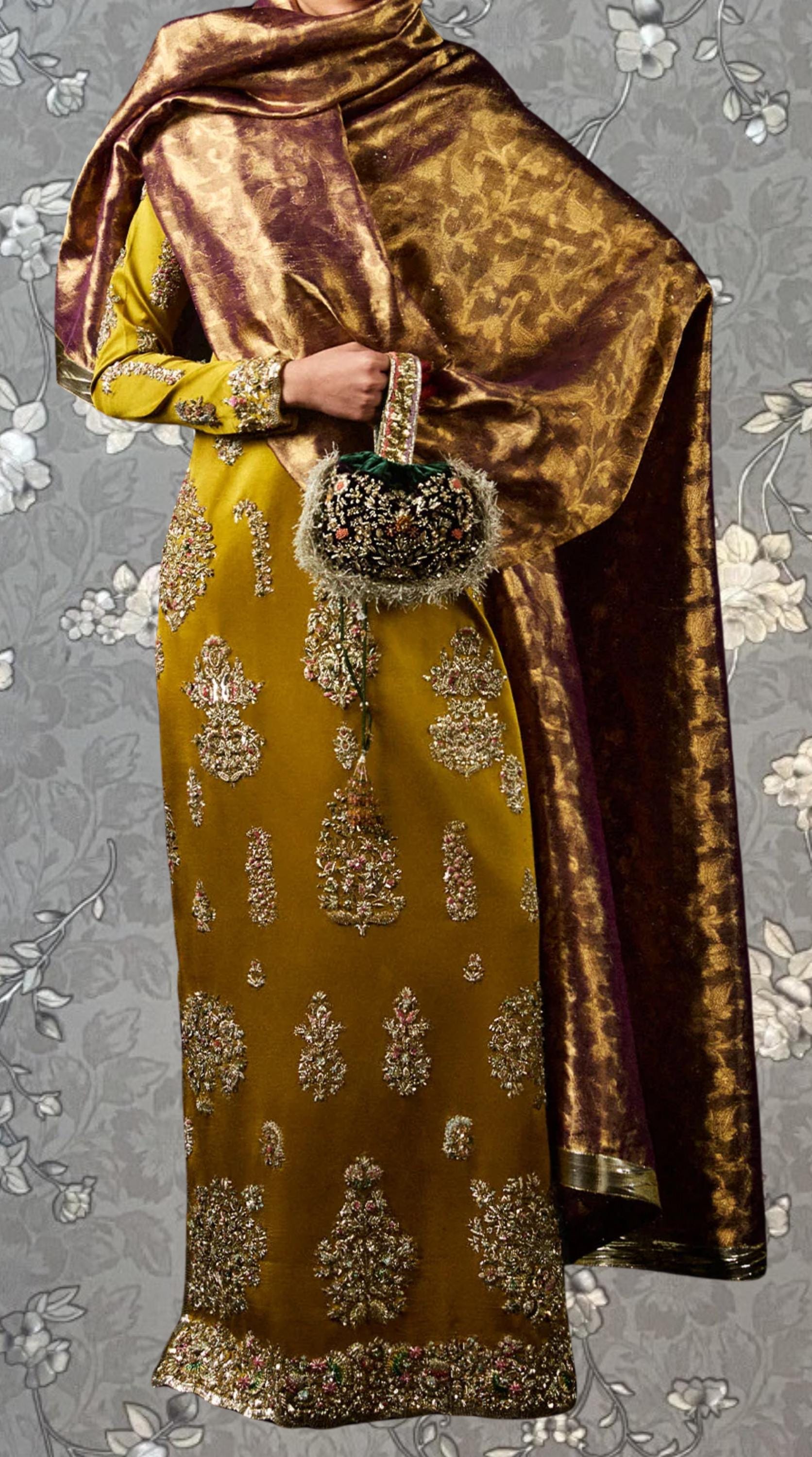 a person wearing a gold and yellow dress with intricate embroidery, holding a small purse, and standing against a gray floral background.