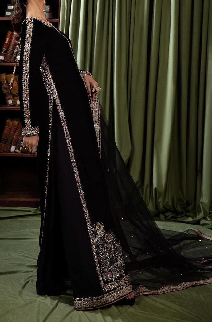 A person wearing a long black dress with intricate embroidery and a flowing black veil stands in front of a green curtain.