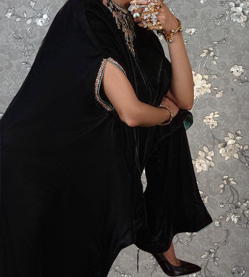 A person wearing a black dress with gold accents and jewelry is standing against a floral background.