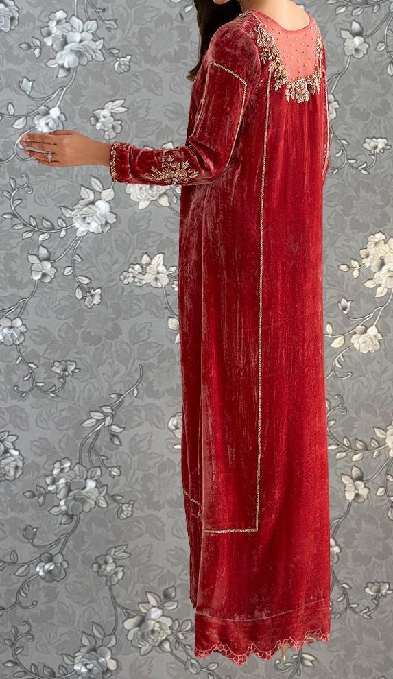 A woman wearing a long red velvet dress with intricate embroidery stands against a gray floral background.