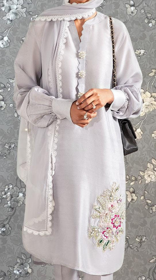 A person wearing a white dress with floral embroidery and a matching white scarf stands against a gray floral background.
