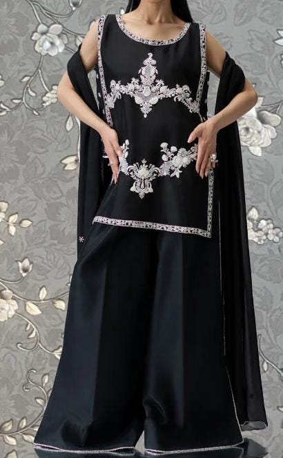 A woman wearing a black dress with white embroidered designs, posing against a gray floral background.
