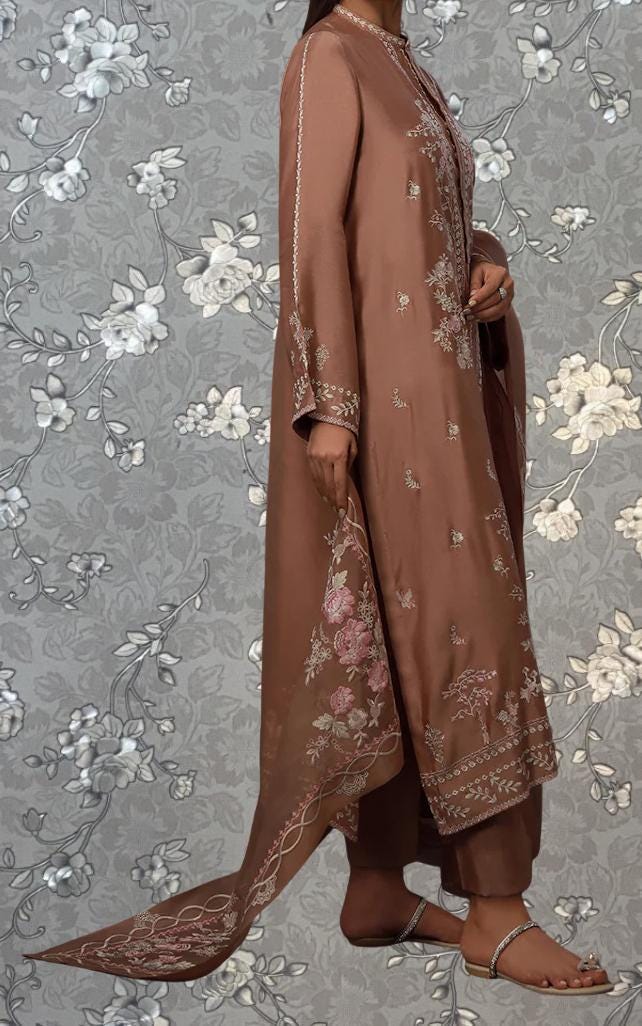 A woman wearing a brown dress with floral embroidery stands against a gray floral background.