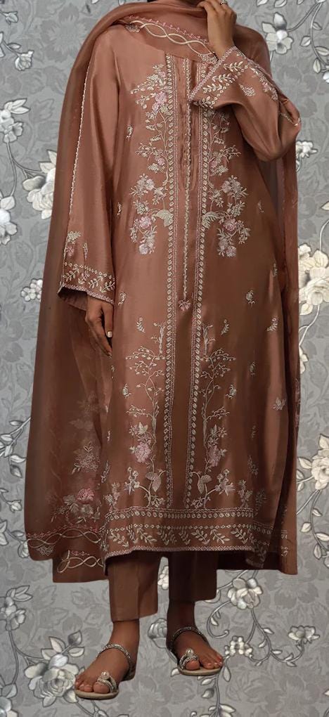 A person wearing a brown, intricately embroidered dress stands against a gray floral background.