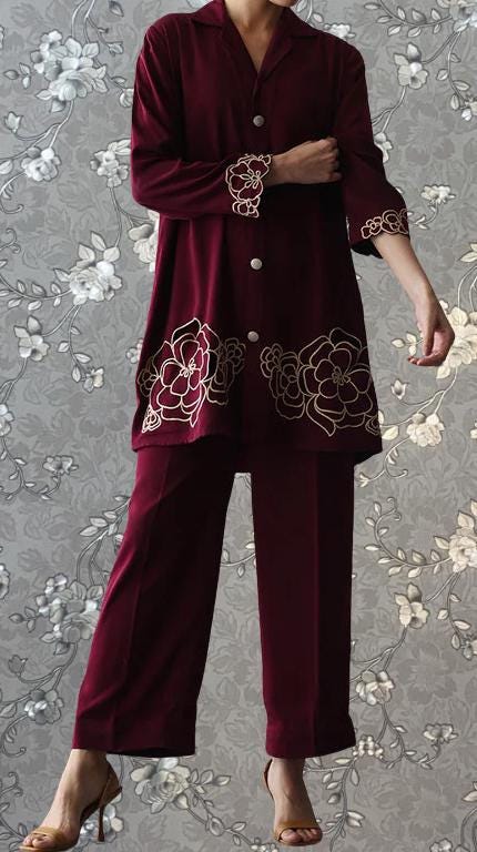 A person wearing a burgundy outfit with gold floral embroidery stands against a gray floral background.