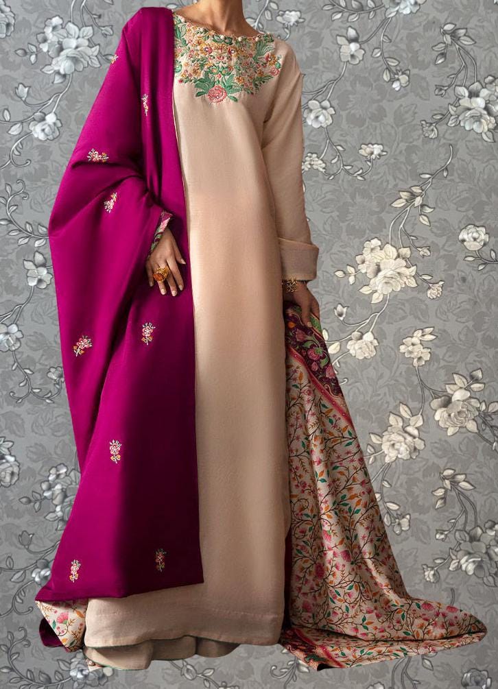 A person wearing a long, beige dress with a pink shawl draped over it, standing against a gray floral background.