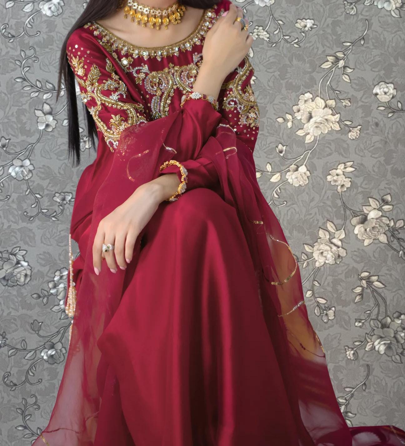 A woman wearing a red and gold dress with intricate embroidery and jewelry poses against a gray floral background.