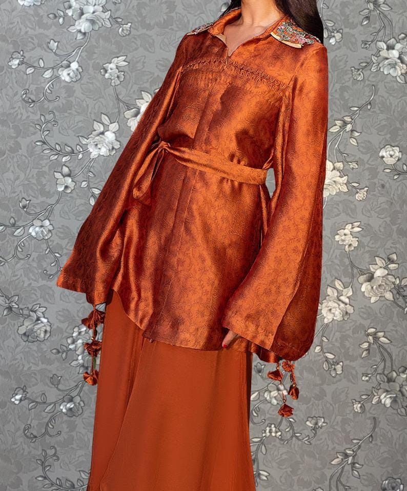 A woman wearing a long orange dress with a high collar and long sleeves, standing against a gray floral background.