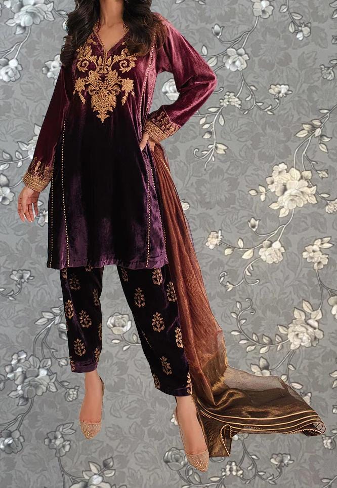 A woman wearing a long, burgundy velvet dress with gold embroidery and gold-colored pants stands against a gray floral background.