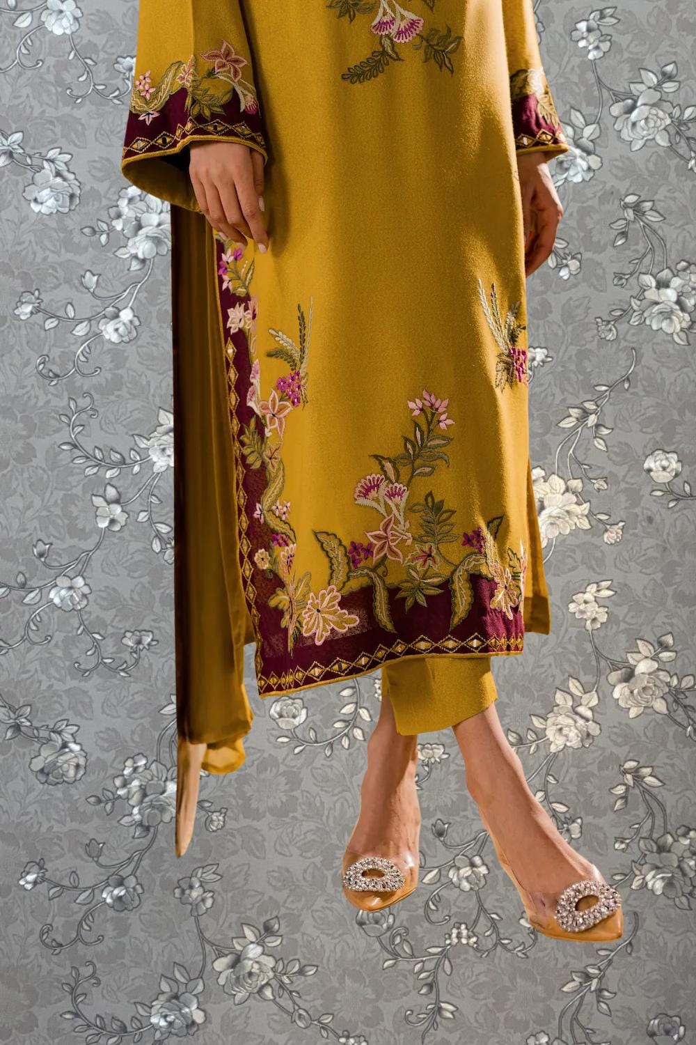 A person wearing a yellow dress with intricate floral embroidery, standing against a gray floral background.