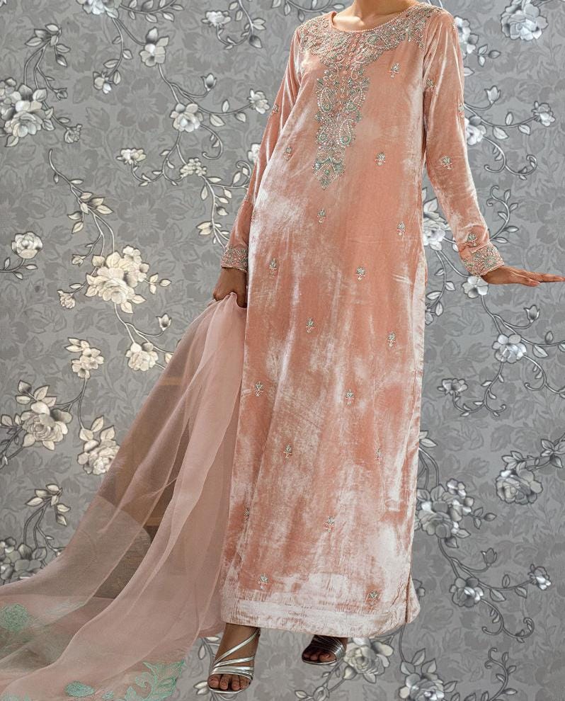 A woman wearing a long, peach-colored dress with intricate embroidery stands against a gray floral background.