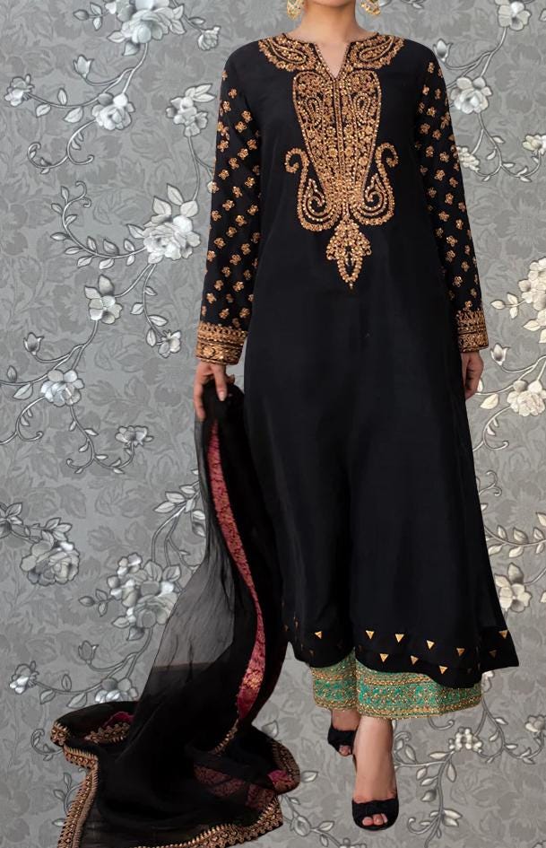 A woman wearing a black and gold dress with intricate embroidery and patterns, standing against a gray floral background.
