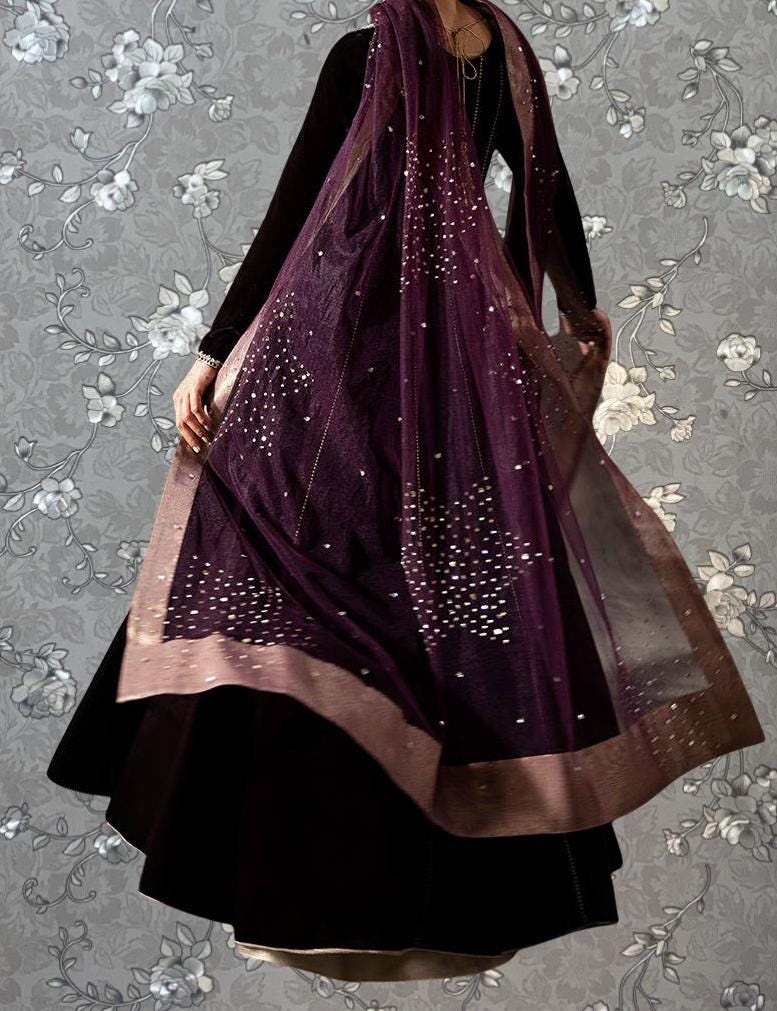 A person wearing a long, purple dress with a sheer overlay and gold embellishments, standing against a gray floral background.