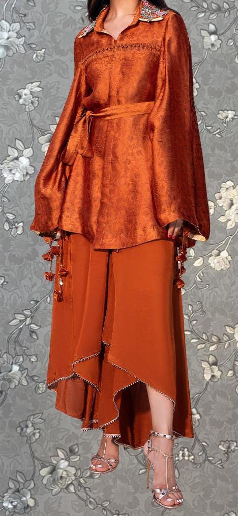 A woman wearing an orange dress with a high collar and long sleeves, standing against a gray floral background.