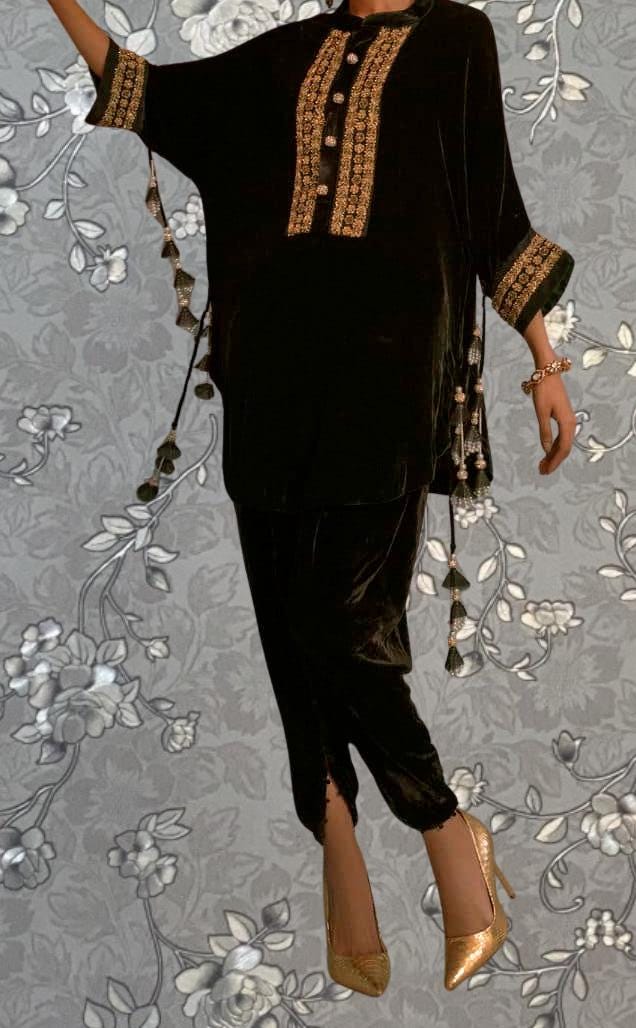 A person wearing a black velvet dress with gold embroidery and tassels, paired with gold high heels, stands against a gray floral background.