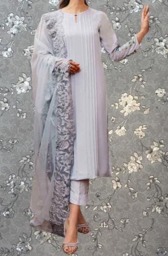A woman wearing a long gray dress with a sheer gray and white patterned shawl draped over her shoulders, standing against a gray floral background.