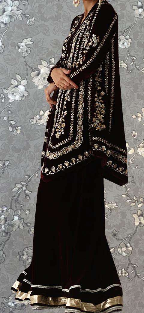 A woman wearing a long, dark dress with intricate gold embroidery stands against a gray floral background.