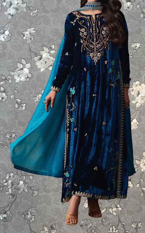 A woman wearing a long, blue velvet dress with intricate embroidery and a matching blue cape.