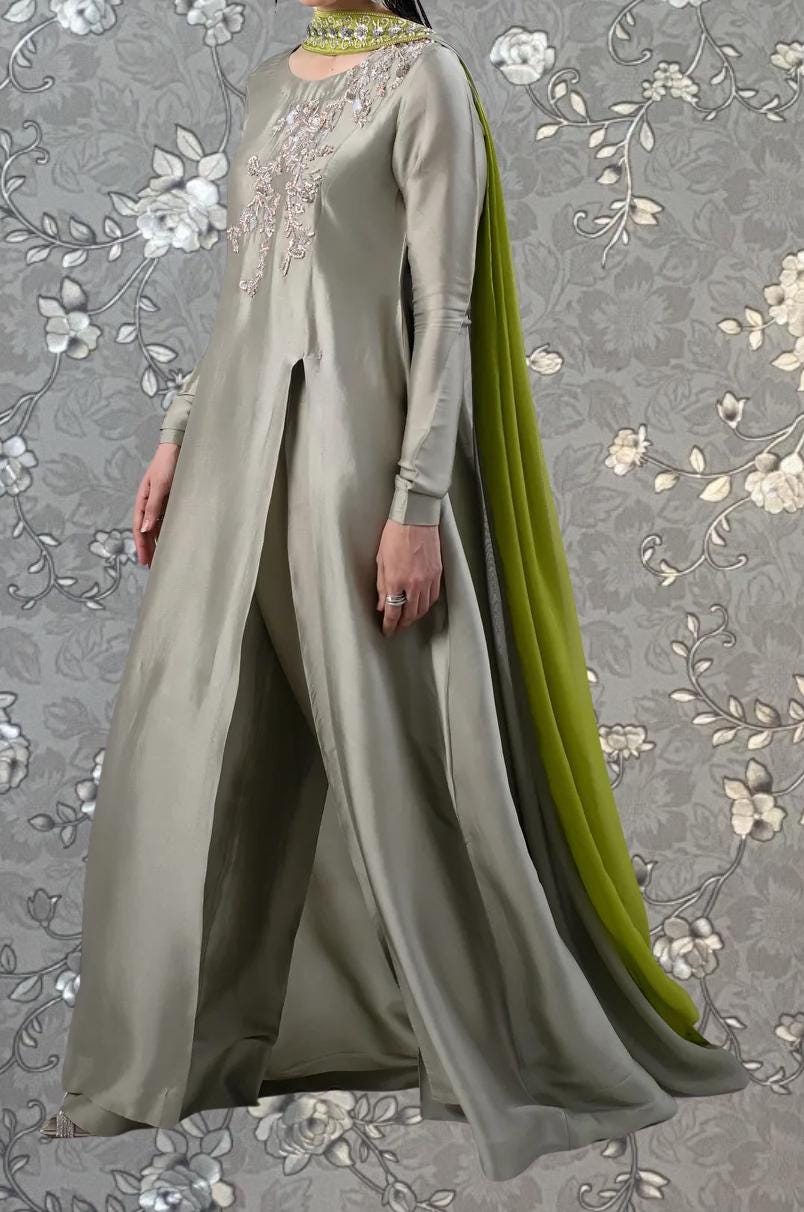 A woman wearing a long, gray dress with a green scarf draped over her shoulders, standing against a gray floral background.
