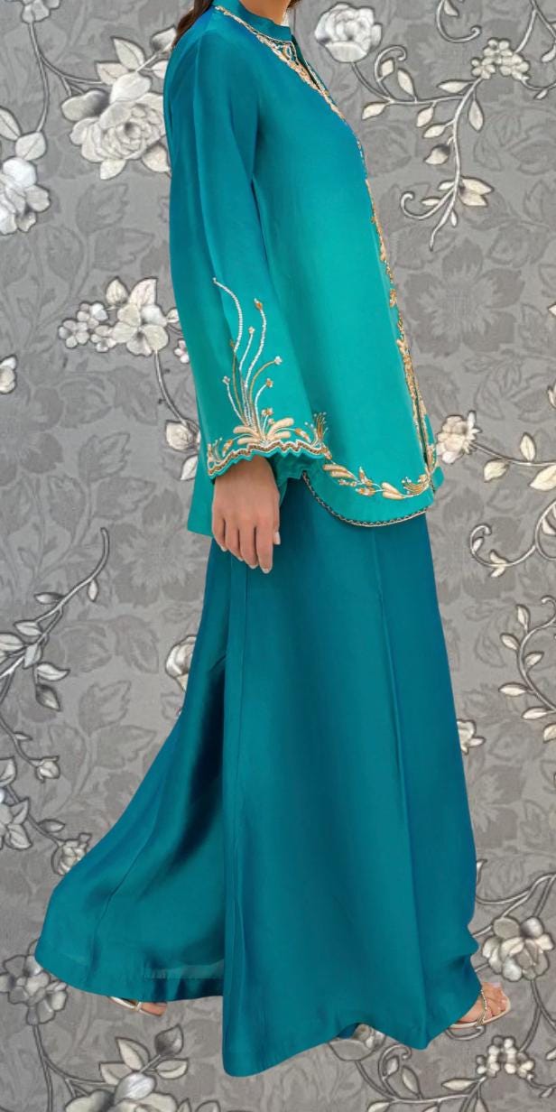 A woman wearing a long, turquoise dress with gold embroidery stands against a gray floral background.
