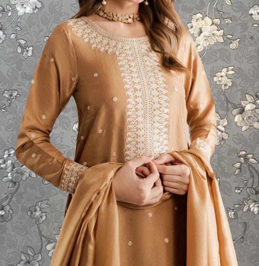 A woman wearing a gold and white dress with intricate embroidery stands against a gray floral background.