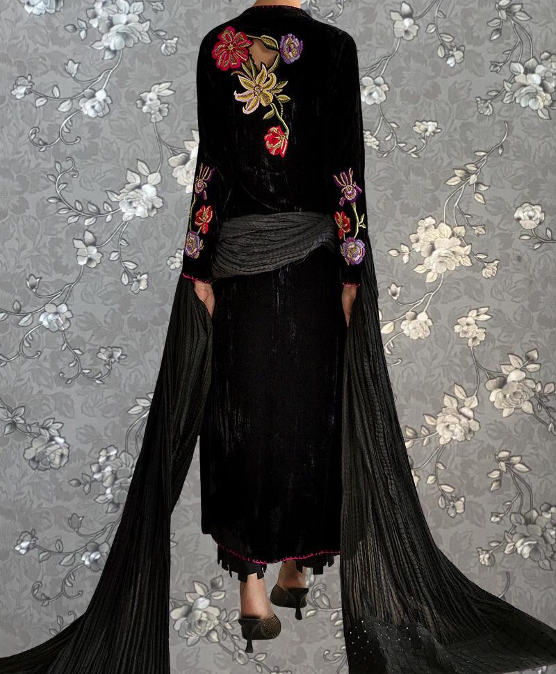 A person wearing a long black dress with floral embroidery on the back, standing against a gray floral background.
