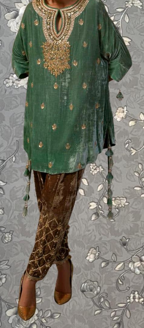 A person wearing a green dress with gold embroidery and tassels, paired with gold and brown patterned pants, standing against a gray floral background.