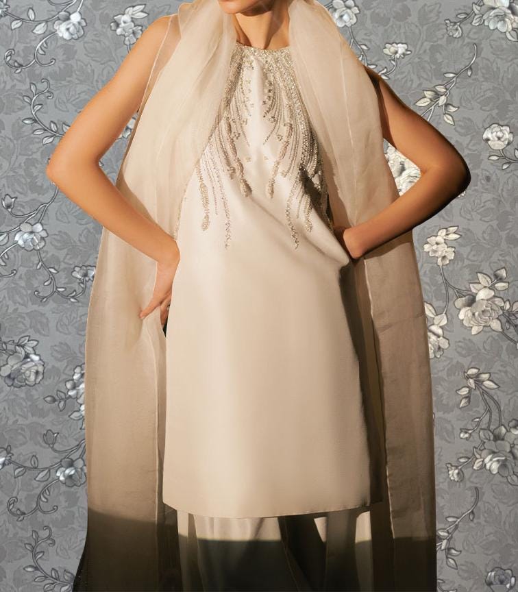 A woman wearing a white dress with a beaded neckline and a sheer white scarf draped over her shoulders, posing against a gray floral background.