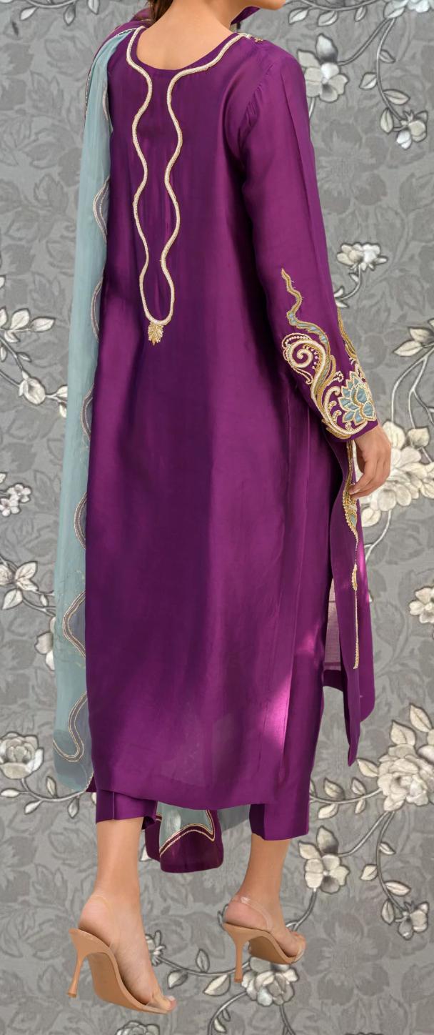 A person wearing a long purple dress with intricate embroidery and gold accents, standing against a gray floral background.