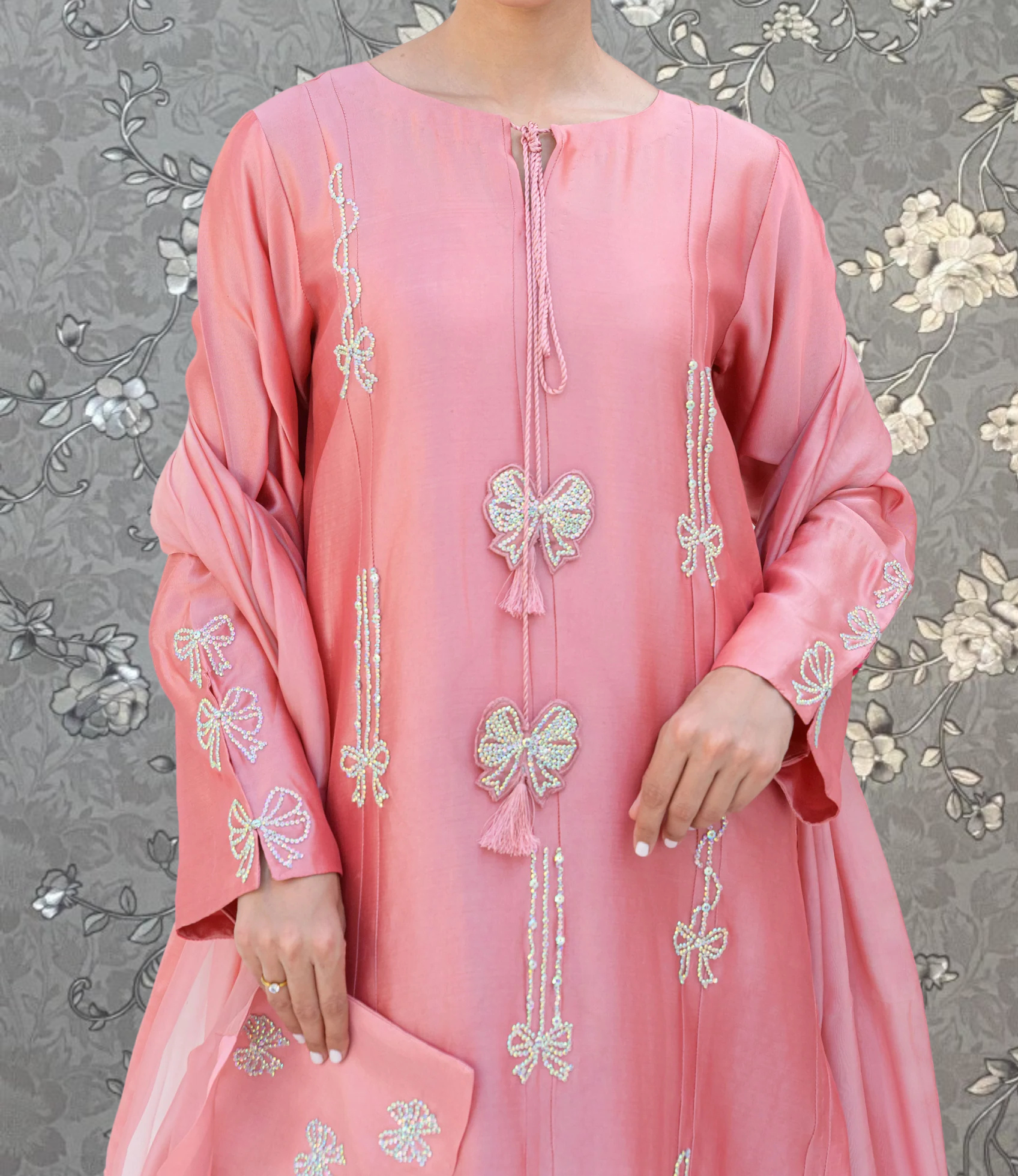 A woman wearing a pink dress with intricate embroidered designs, including bows and flowers, stands against a gray floral background.