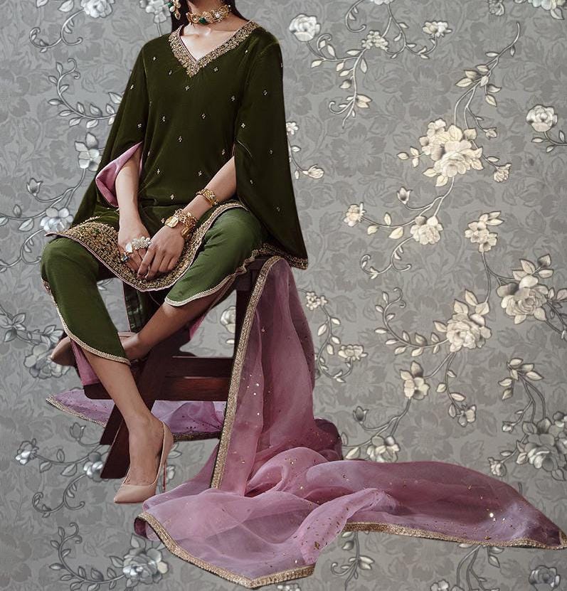 A woman wearing a green and pink dress with a pink shawl is sitting on a wooden chair against a gray floral background.