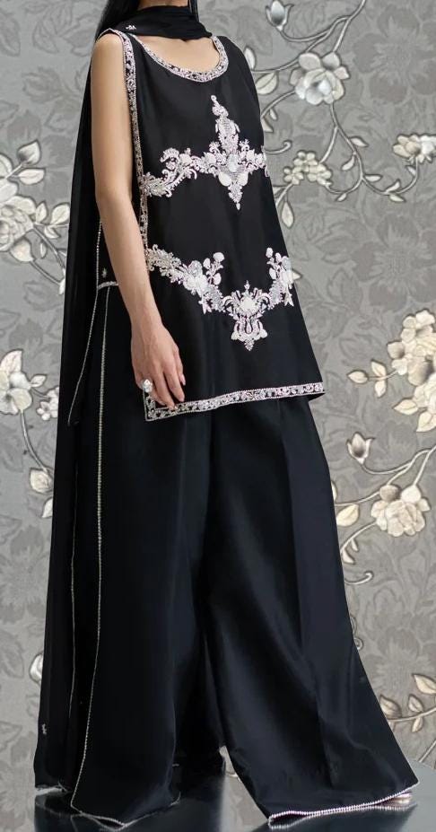 A person wearing a black dress with white embroidered designs, standing against a gray floral background.