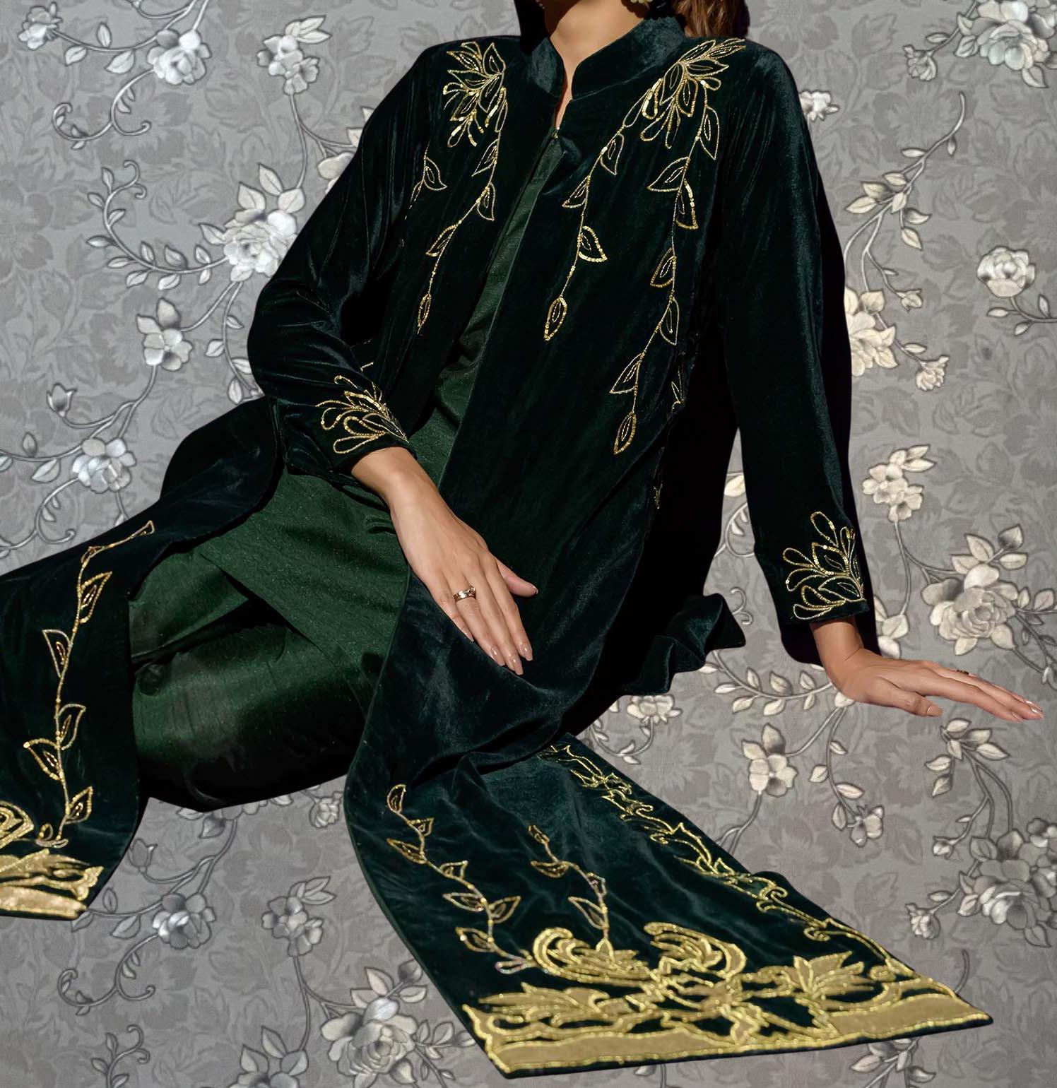 A person wearing a long, green velvet coat with gold embroidered designs, sitting on a gray floral background.